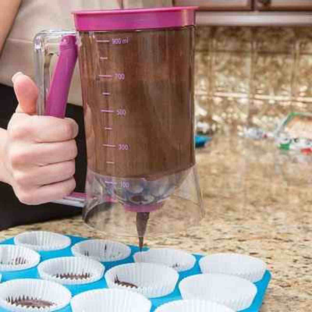Entonnoir à piston transparent rempli de pâte foncée, dosant avec précision dans des caissettes à muffins blanches sur un plan de travail en granit.