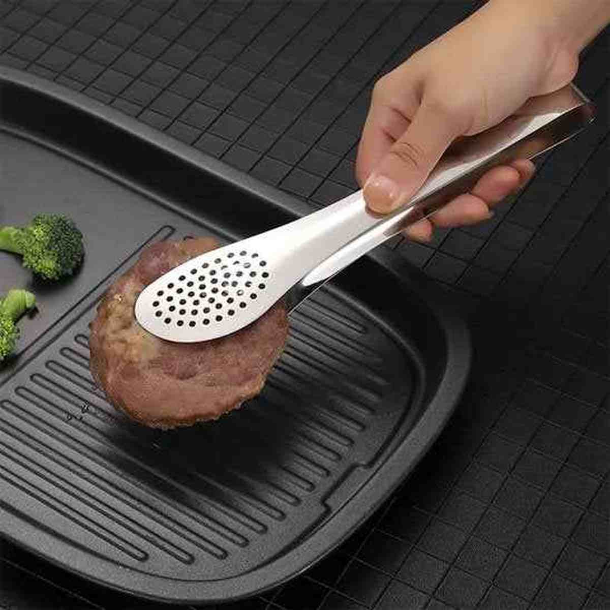 Pince de cuisine en acier inoxydable avec tête perforée saisissant un steak sur une poêle grill avec des légumes