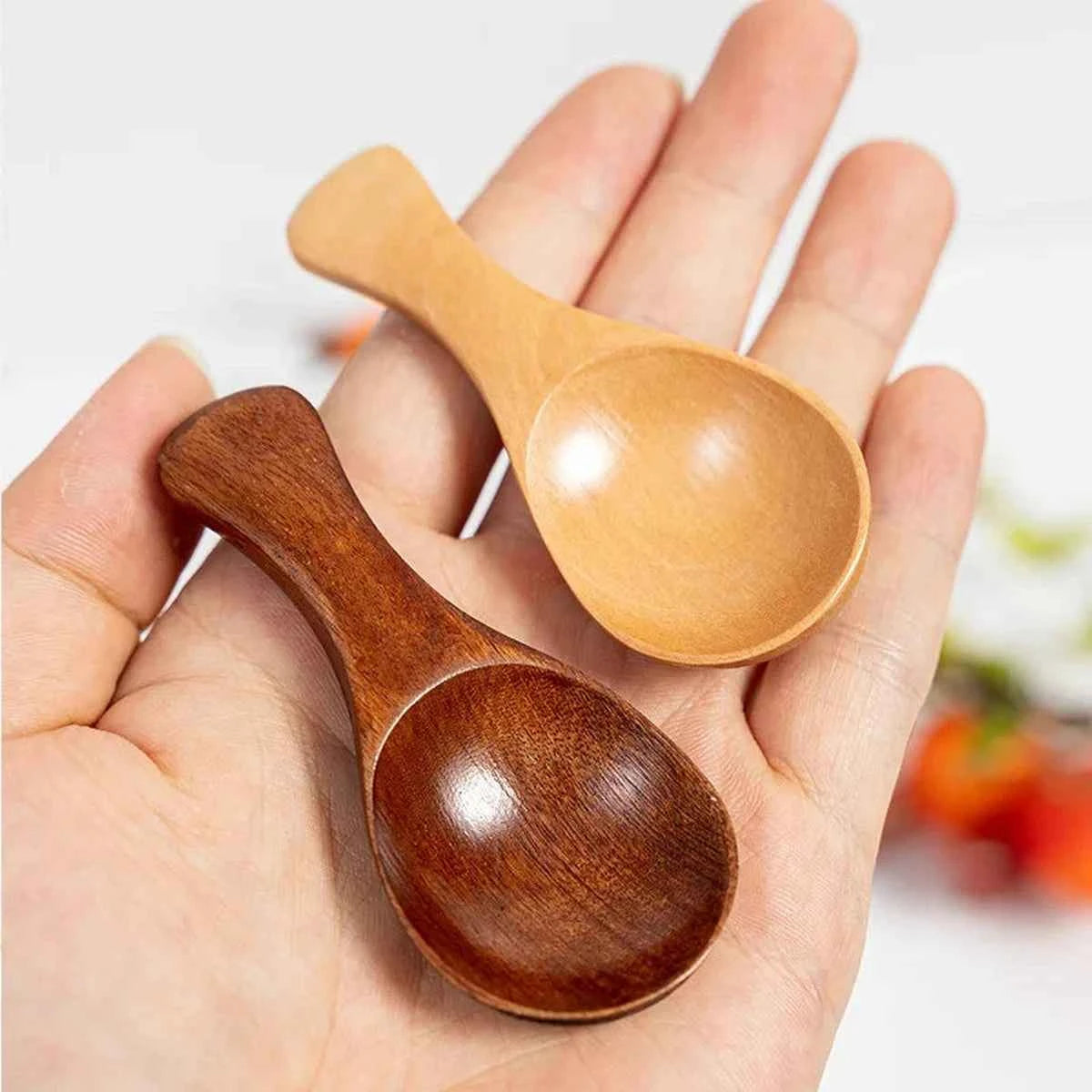 Main tenant deux mini cuillères de dosage en bois pour montrer la taille compacte et les deux finitions disponibles : bois naturel et marron foncé