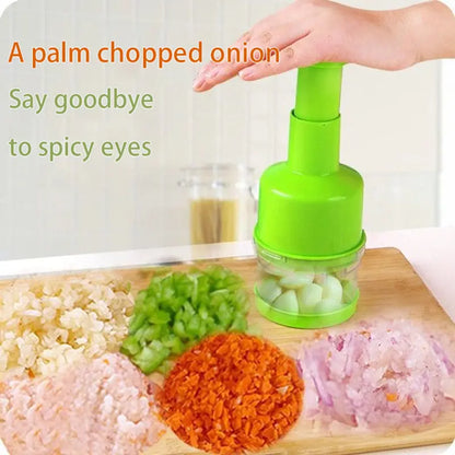 Hachoir manuel à pression vert utilisé pour couper des oignons et de l'ail sur une planche en bois dans une cuisine lumineuse.