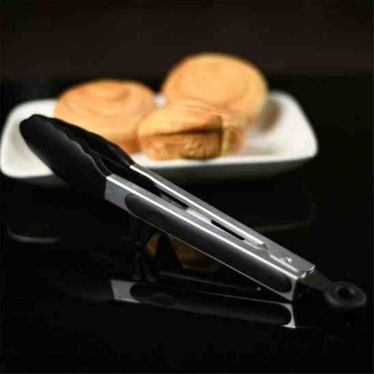 Pince de cuisine en acier inoxydable et silicone noir posée sur une table à côté d'une assiette de petits pains dorés.