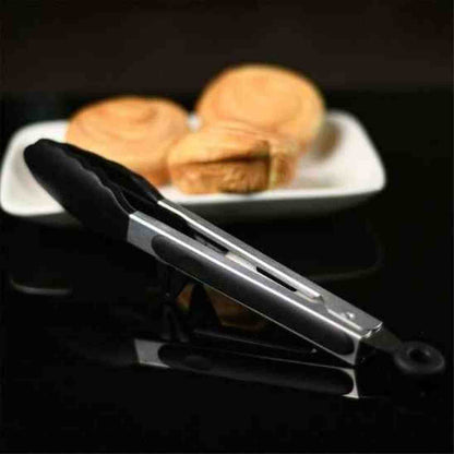 Pince de cuisine en acier inoxydable et silicone noir posée sur une table à côté d'une assiette de petits pains dorés.