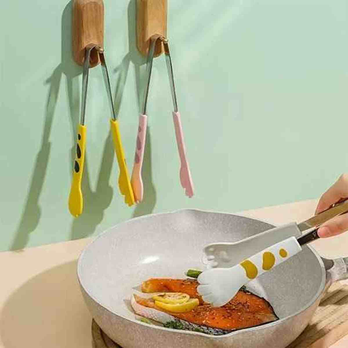 Pince de cuisine en forme de patte de chat blanc avec taches brunes, utilisée pour retourner un filet de poisson dans une poêle, idéale pour les surfaces délicates.