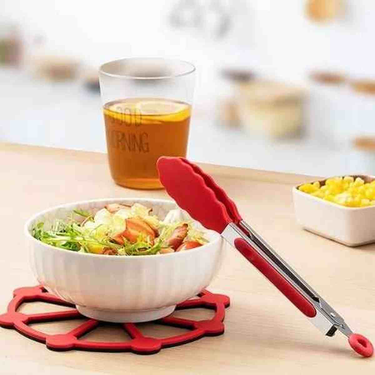 Pince de cuisine en silicone rouge posée à côté d'un bol de salade fraîche et d'une boisson, illustrant un usage quotidien et convivial.