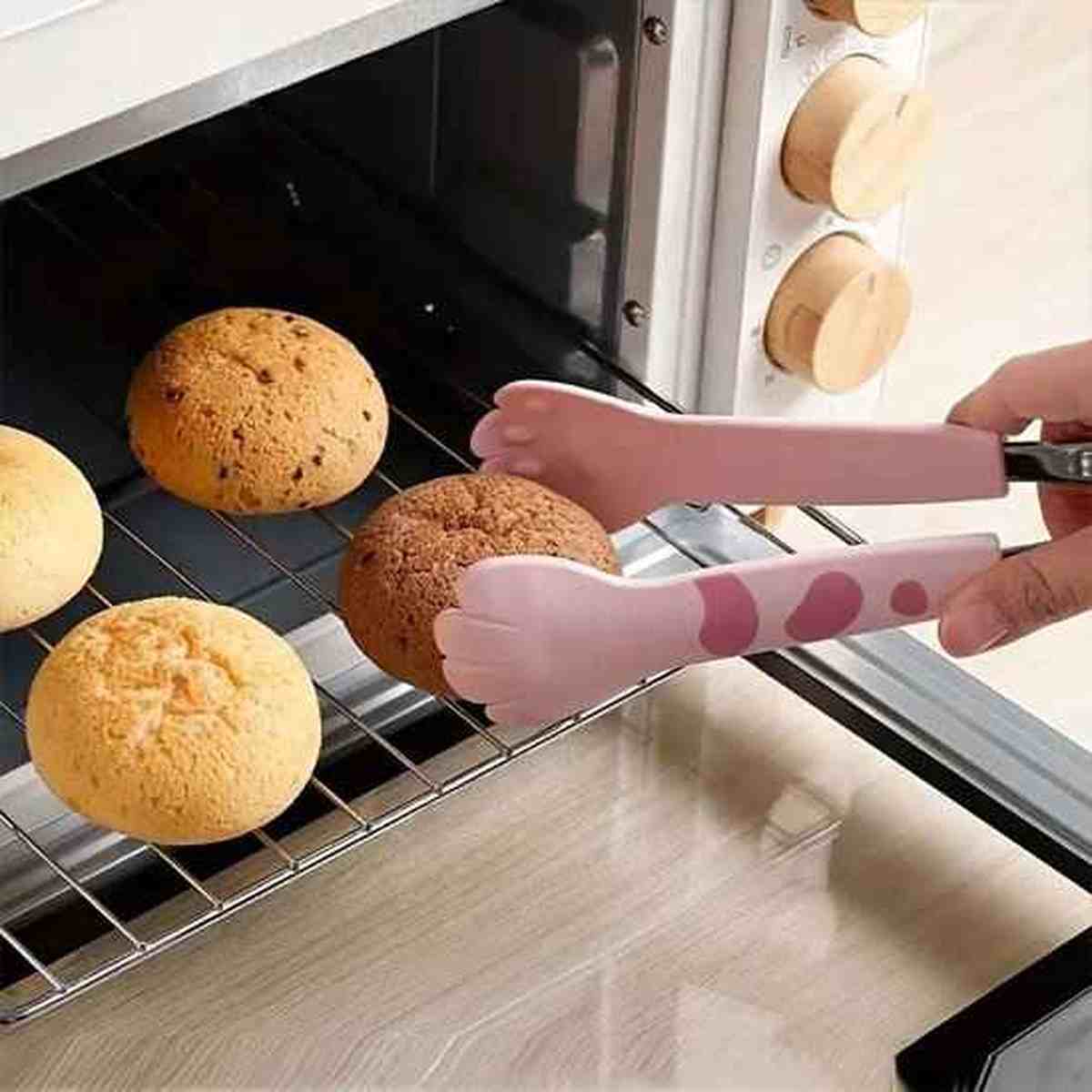 Pince de cuisine ergonomique en silicone rose en forme de patte de chat saisissant un pain chaud dans le four.