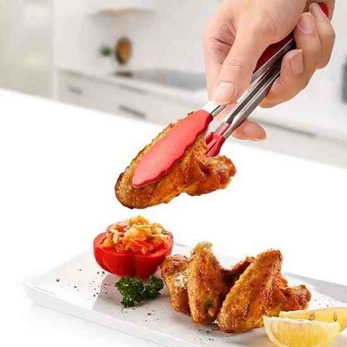 Main tenant une pince en silicone rouge pour servir des ailes de poulet grillées croustillantes sur une assiette de présentation.