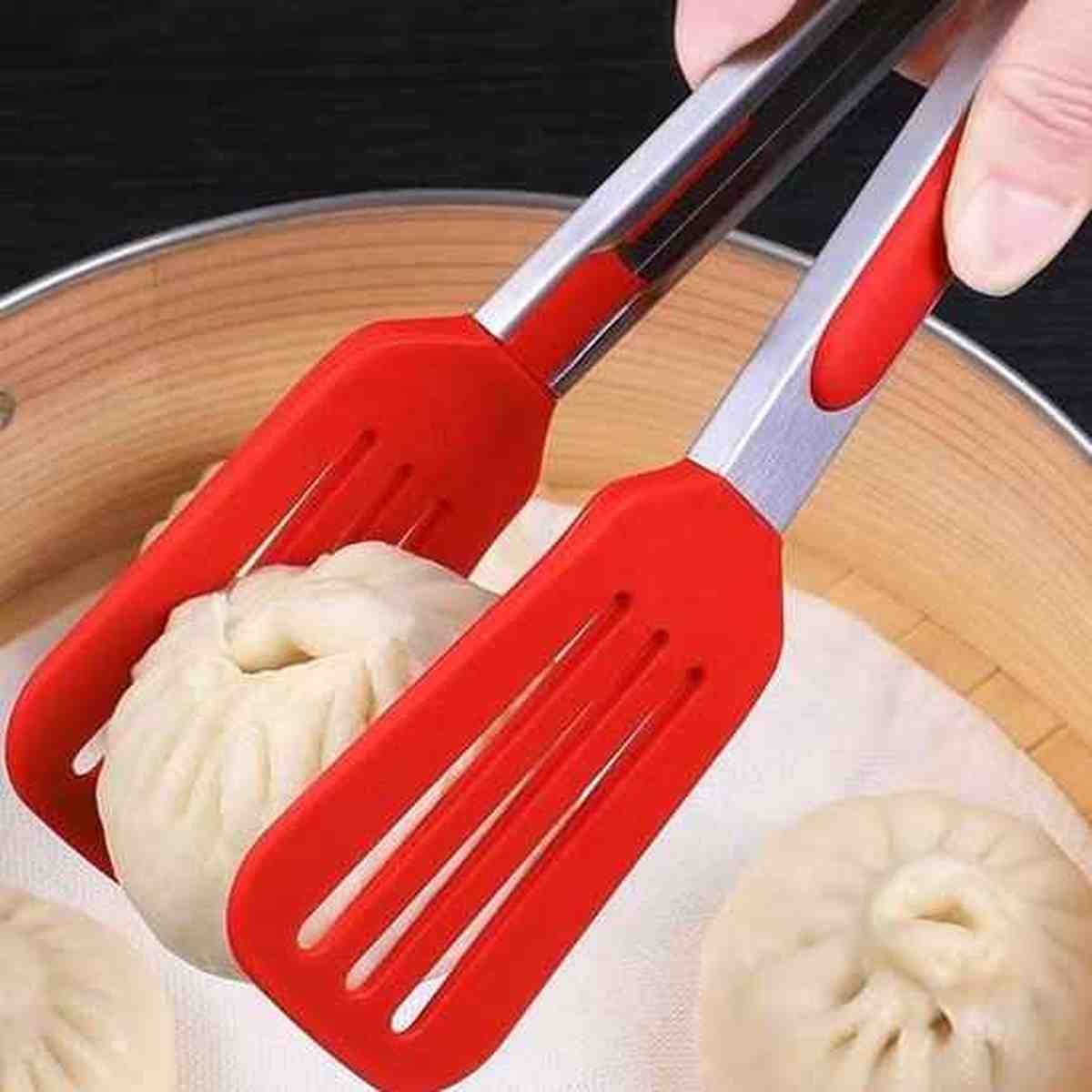 Pince de cuisine en silicone rouge saisissant avec précision un ravioli vapeur dans un panier en bambou.