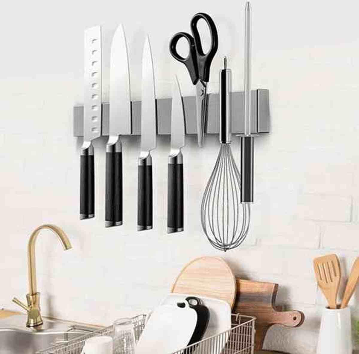 Barre magnétique en acier inoxydable fixée au mur d'une cuisine blanche, tenant plusieurs couteaux de chef, des ciseaux et un fouet.
