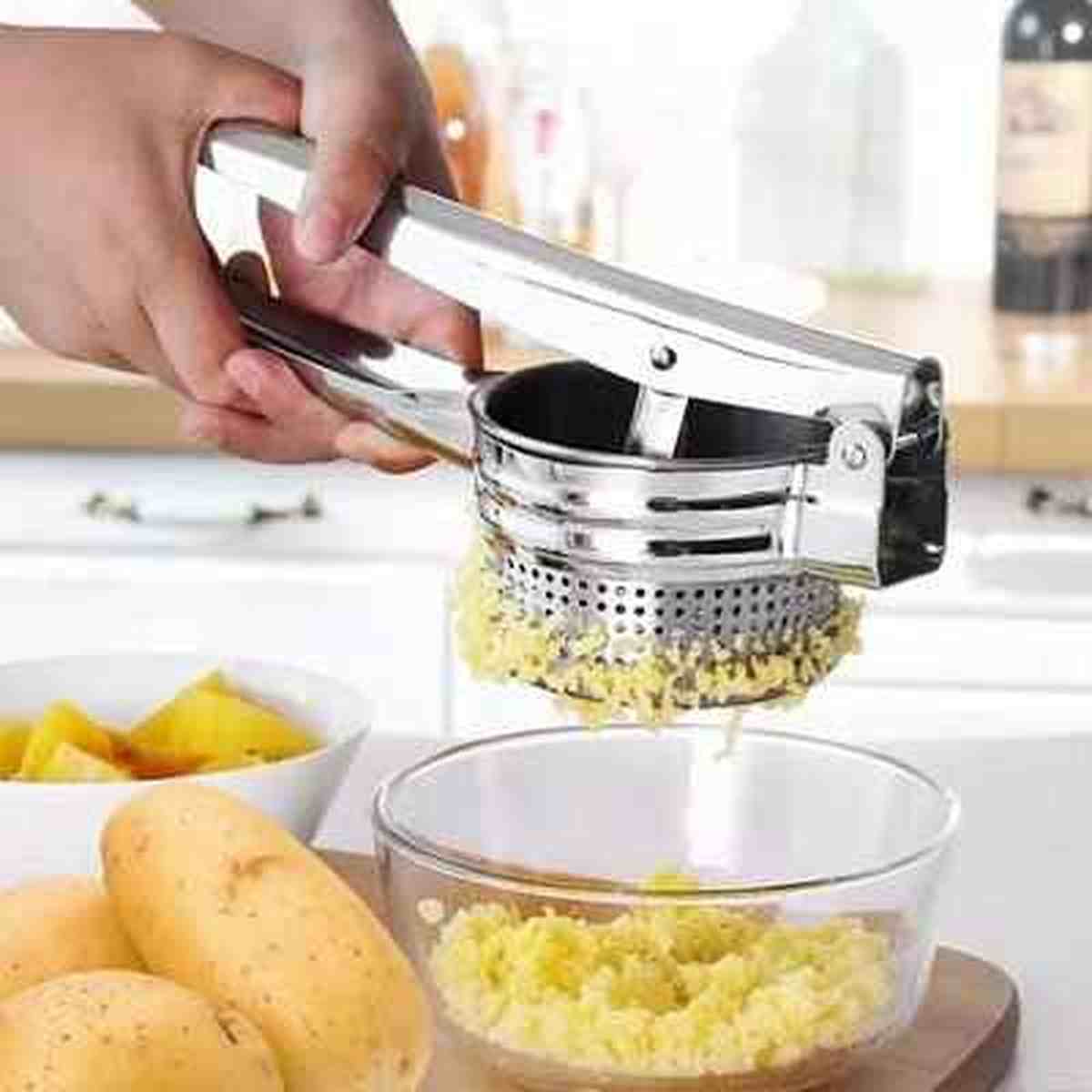Utilisation du presse-purée manuel ergonomique en acier inoxydable pour écraser des pommes de terre au-dessus d'un bol en verre.