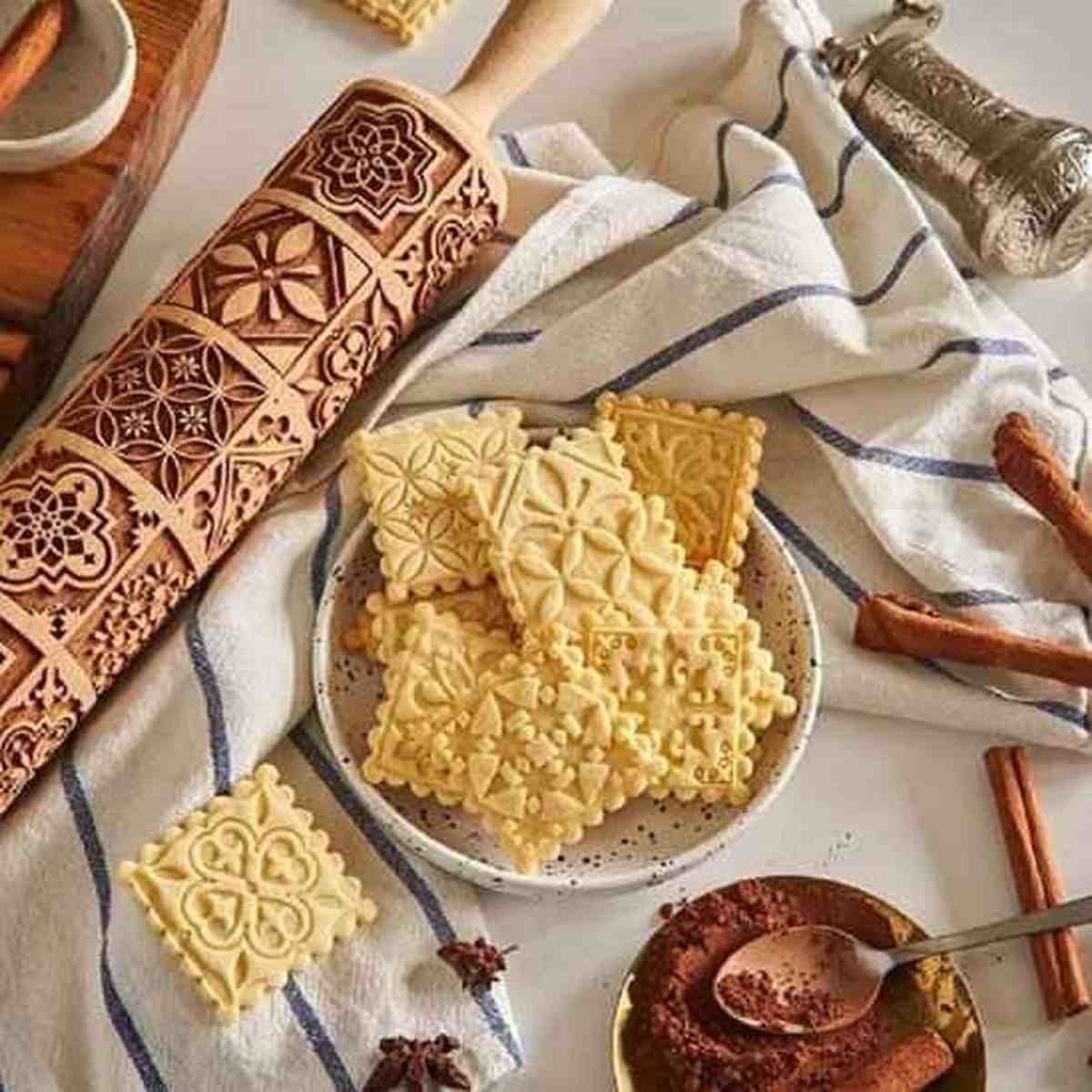 Biscuits artisanaux aux motifs géométriques et floraux présentés dans une assiette à côté du rouleau à pâtisserie en bois gravé Artisan Pro.